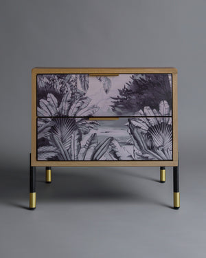 A wooden bedside pedestal with two drawers featuring a monochromatic forest design and brass-accented legs.