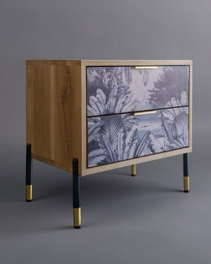 A wooden bedside pedestal with two drawers featuring a monochromatic forest design and brass-accented legs.