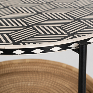 Close-up of a round table with a black and off-white geometric inlay pattern on the tabletop and a black metal leg, with a woven chair partially visible underneath.