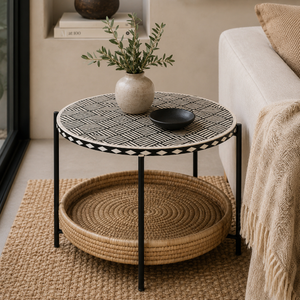 A round two-tier side table in a styled living room. The top table has a black and white geometric patterned surface with a black and white border, supported by slim black metal legs.