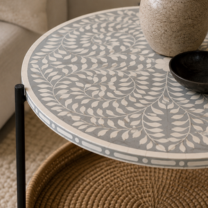 A round nesting side table with a gray and cream leaf patterned top, a black metal leg, and a woven brown base, styled in a cozy living room setting.