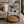 A two-tier round coffee table in a styled living room. The top tier has a white and gray bohemian-style geometric pattern, supported by black metal legs, while the bottom tier features a natural woven rattan tray.