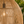 A close-up of a large woven rattan pendant lamp shade in a warm natural tan color, with a glowing light bulb visible inside and an outdoor garden background.