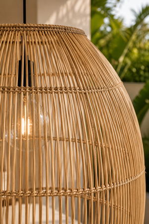 A close-up of a large woven rattan pendant lamp shade in a warm natural tan color, with a glowing light bulb visible inside and an outdoor garden background.