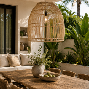 A large woven rattan pendant light hangs over a rustic wooden dining table in an outdoor patio setting with chairs, a sofa, and lush green plants.