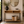 A modern console table with a light wood top, black metal frame, and woven lower shelf, styled in a bright entryway with a mirror, plant, books, and basket.