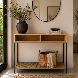 A modern console table with a light wood top, black metal frame, and woven lower shelf, styled in a bright entryway with a mirror, plant, books, and basket.