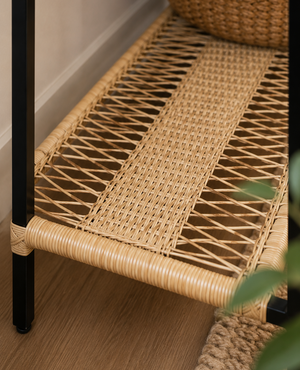 Close-up of a woven rattan or wicker shelf with a black metal frame, placed on a wood floor next to a woven basket and a plant.