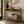 A light wood console table with open upper shelves and a woven lower shelf, styled in a bright entryway with a black metal frame. The table holds a dark ceramic vase with greenery, a bowl, stacked books, and a woven basket with a cream throw blanket.