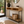 A modern console table with a light wood top, black metal frame, and woven lower shelf, styled in a bright entryway with a mirror, plant, books, and basket.