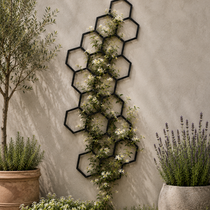 A black metal hexagonal trellis wall decor piece with climbing green vines and small white flowers, mounted on a textured beige stucco wall in a garden setting with potted plants.