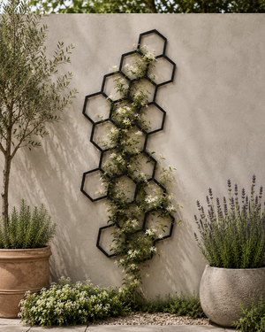 A black metal hexagonal trellis wall decor piece with climbing green vines and small white flowers, mounted on a textured beige stucco wall in a garden setting with potted plants.