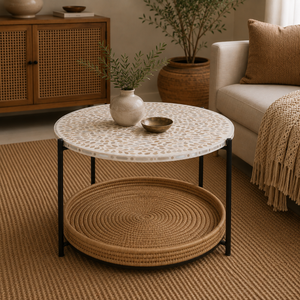 A round two-tier coffee table in a cozy living room. The top table has a white and beige mosaic tile surface with black metal legs, and the bottom tier is a round woven rattan tray.